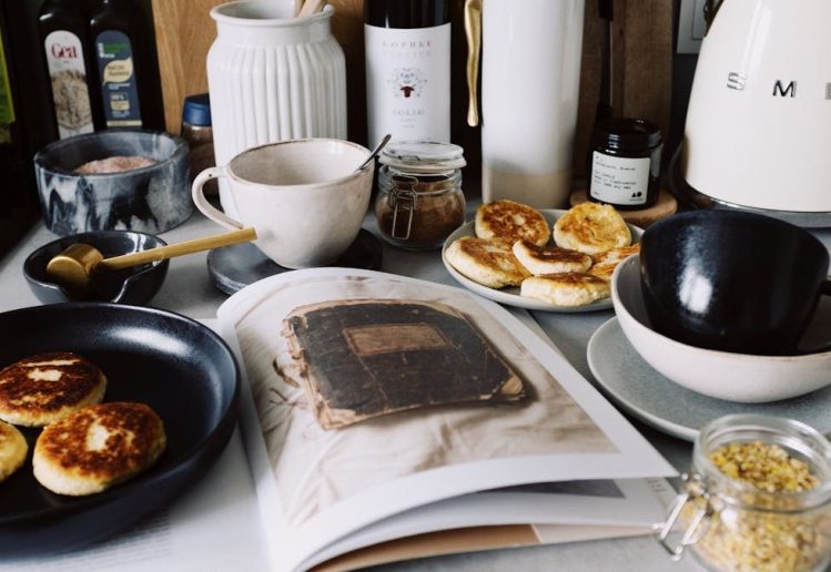 One portion of homemade cheese pancakes in frying pan and another on plate locating near opened stylish magazine among kitchen utensils and appliances in bright kitchen at home