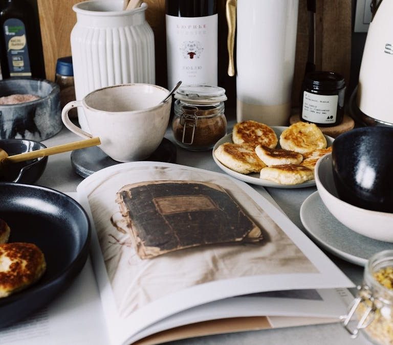 One portion of homemade cheese pancakes in frying pan and another on plate locating near opened stylish magazine among kitchen utensils and appliances in bright kitchen at home